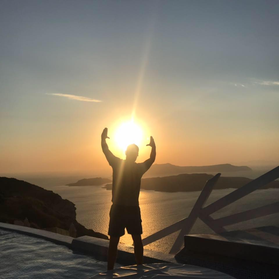 A traveller raising both arms to frame the setting sun over the caldera in Santorini